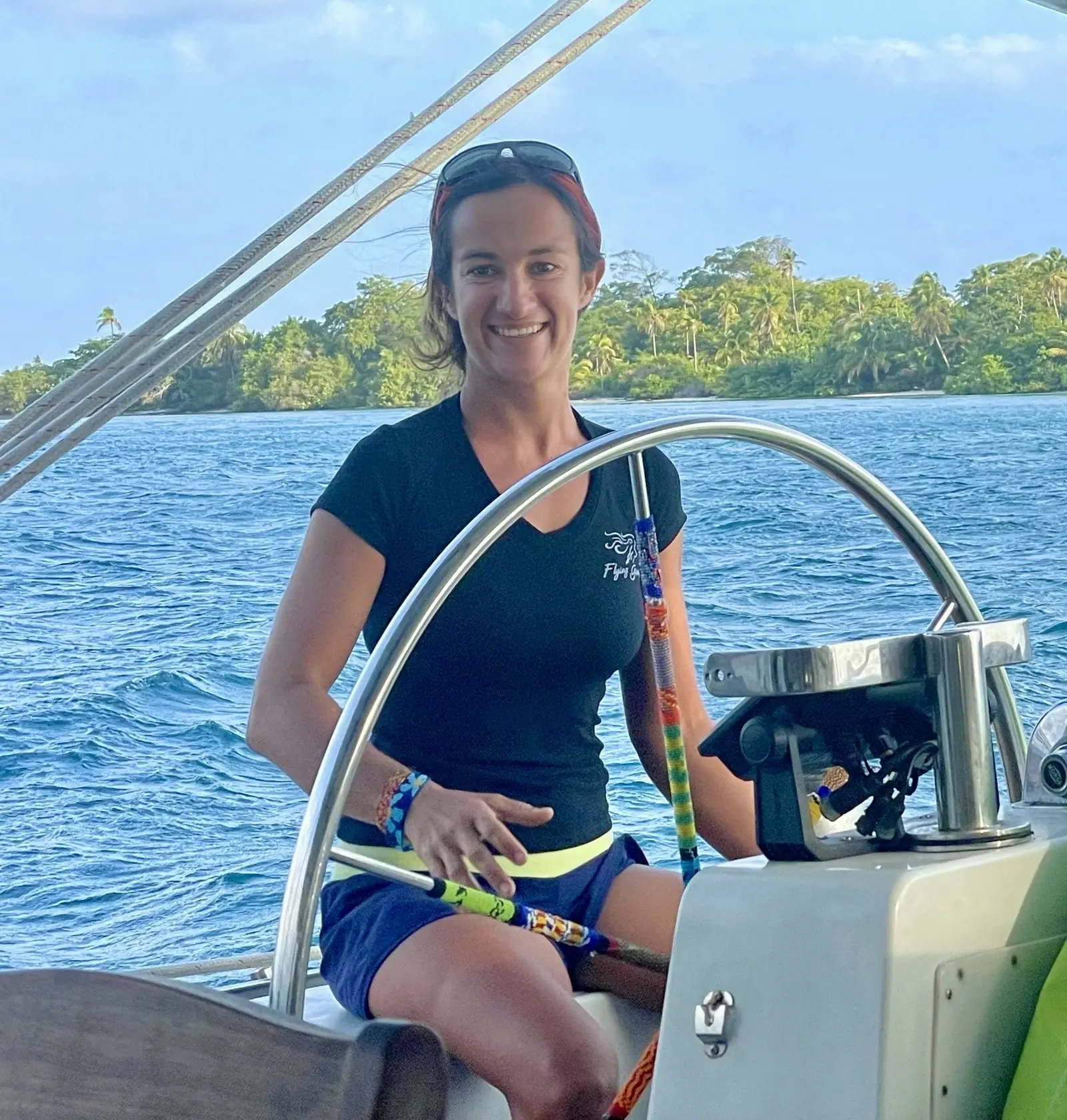 The captain at the wheel of the Lagoon 55 in a branded crew shirt, palm shoreline behind