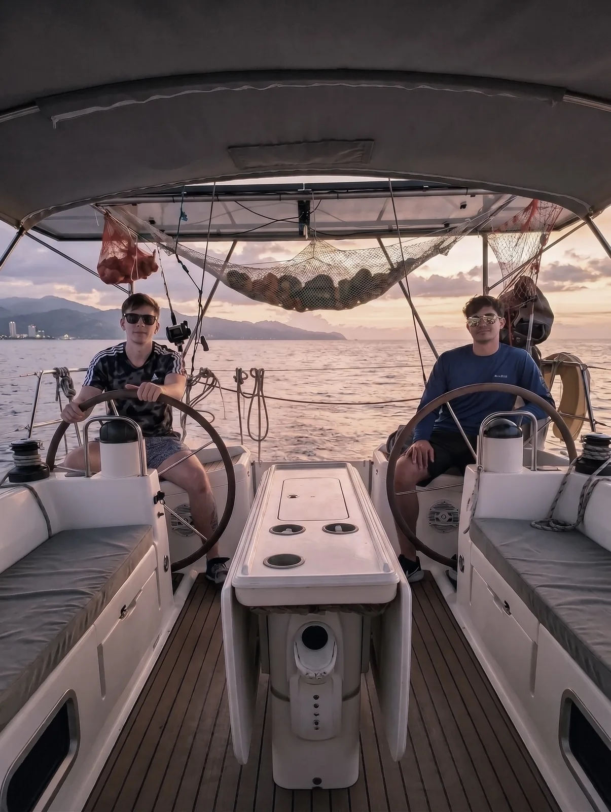 Two people at the twin wheels of the Jeanneau 45.2 at sunset, under the bimini
