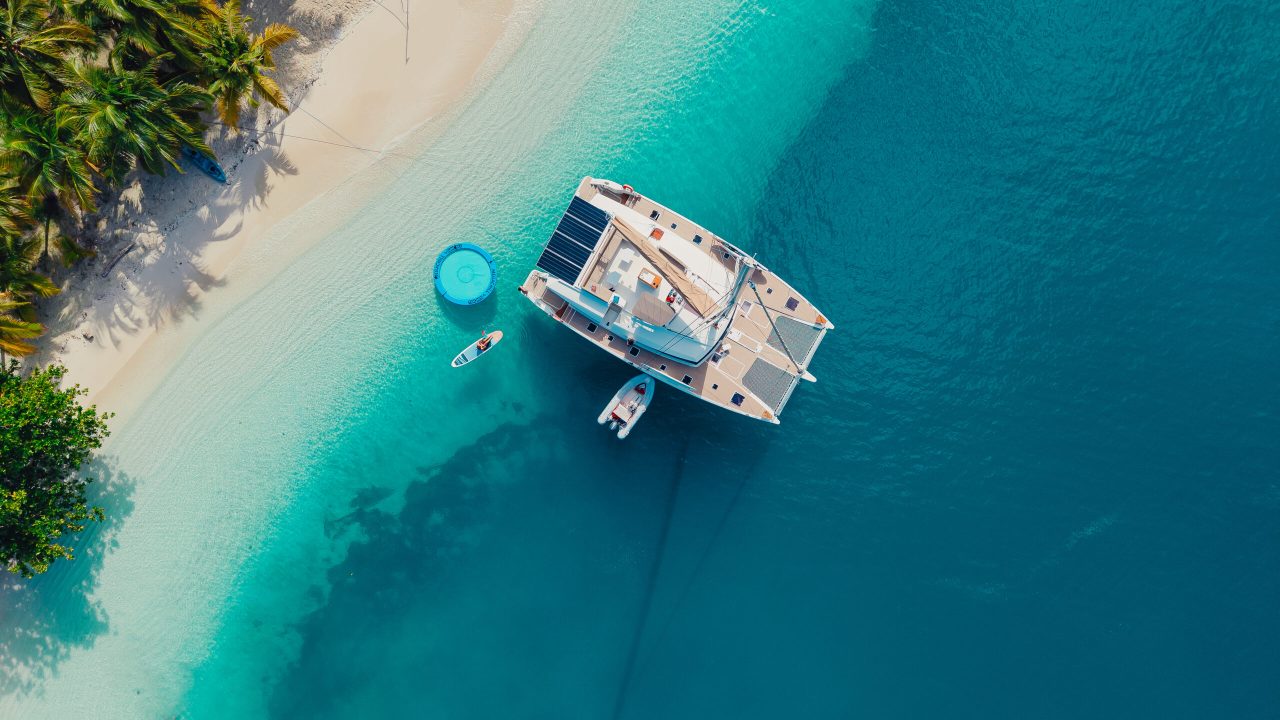 Aerial view of a luxury catamaran anchored near a tropical beach in San Blas, Panama, with clear blue waters and palm trees.